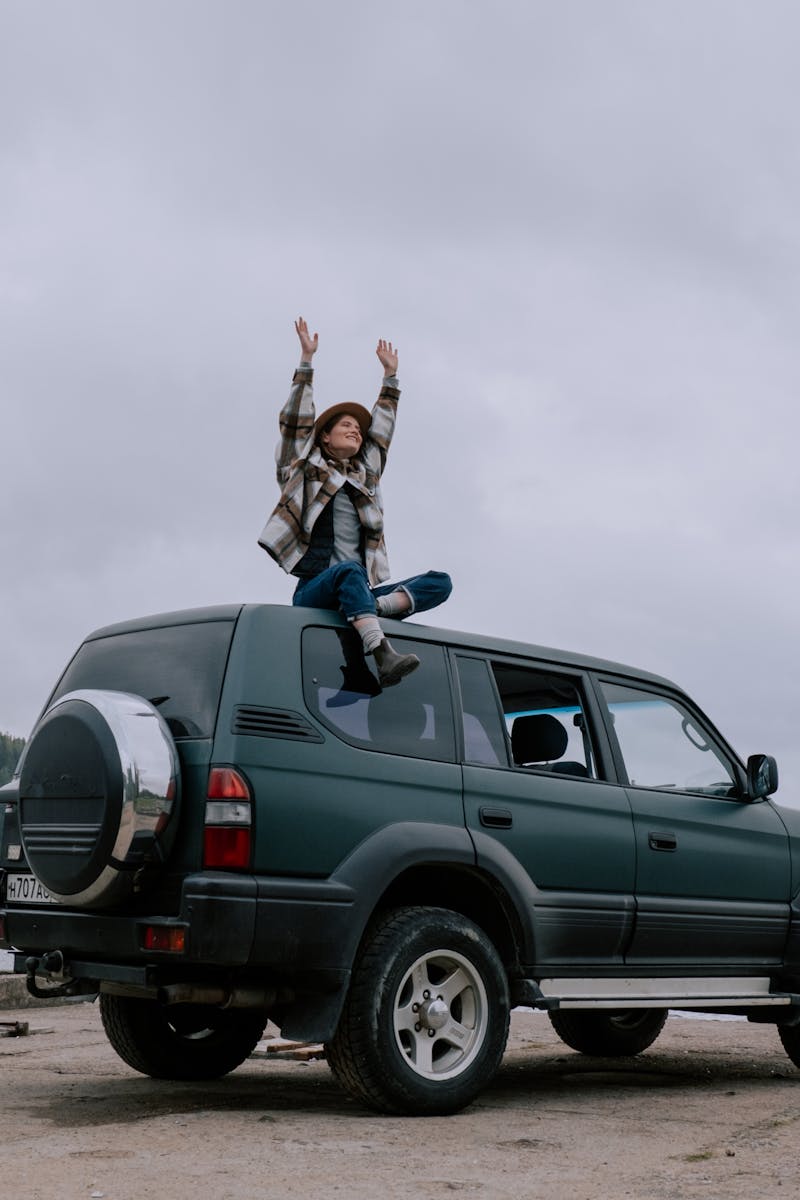 A woman sits on top of an SUV with arms raised in excitement by a scenic lake.